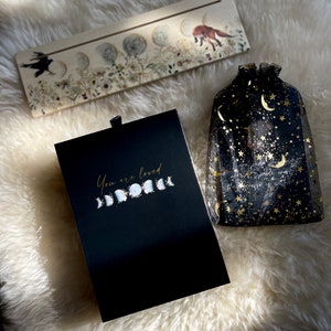 May include: A black box with the text "You are loved" and a gold moon phase design. The box is sitting on a white fluffy surface next to a wooden sign with a moon phase design and a black mesh bag with gold stars and moons.