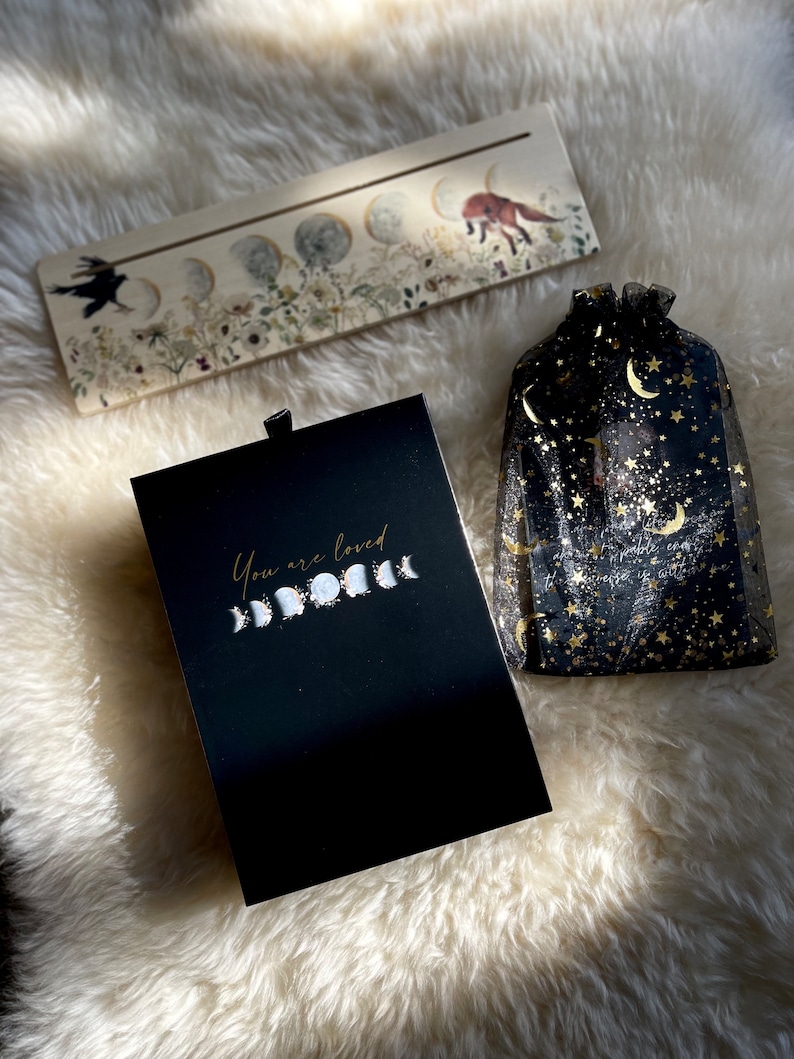 May include: A black box with the text "You are loved" and a gold moon phase design. The box is sitting on a white fluffy surface next to a wooden sign with a moon phase design and a black mesh bag with gold stars and moons.