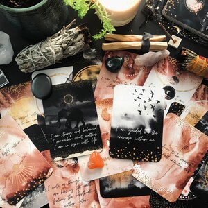 May include: A set of affirmation cards with watercolor backgrounds and gold accents. The cards feature positive messages such as "I am strong and balanced" and "I am guided by the universe within me".