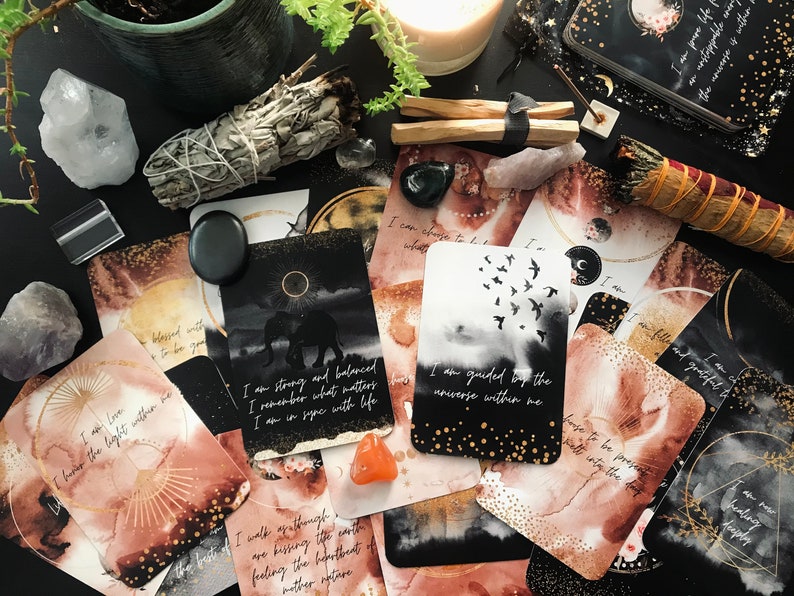 May include: A set of affirmation cards with watercolor backgrounds and gold accents. The cards feature positive messages such as "I am strong and balanced" and "I am guided by the universe within me".