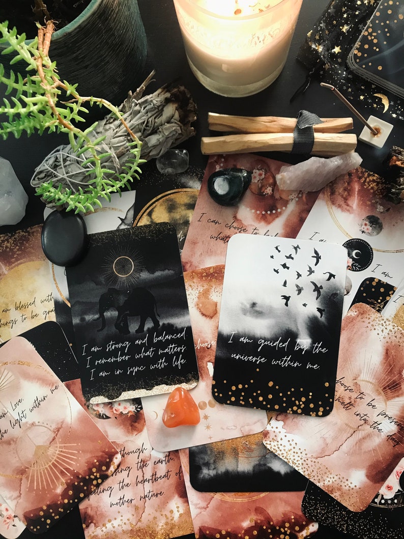 May include: A set of oracle cards with gold and black accents and watercolor backgrounds. The cards feature various illustrations and text, including "I am strong and balanced", "I remember what matters", "I am in sync with life", "I am guided by the universe within me", and "I can choose to be what I want".