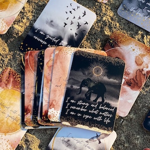 May include: A set of affirmation cards with watercolor backgrounds and gold accents. The cards feature positive messages such as "I am guided by the universe" and "I am strong and balanced".