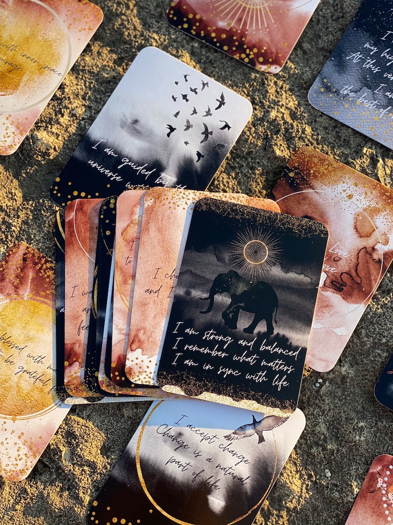 May include: A set of affirmation cards with watercolor backgrounds and gold accents. The cards feature positive messages such as "I am guided by the universe" and "I am strong and balanced".