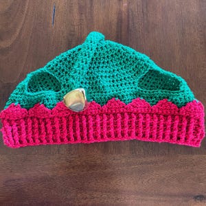 May include: A green and pink crocheted hat with a bell attached. The hat has a scalloped edge and is made with a soft yarn.