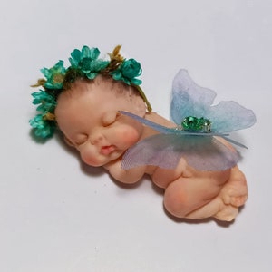 Puede incluir: Una pequeña muñeca de bebé durmiendo con una corona de flores verdes y un ala de mariposa azul. La muñeca está hecha de un material suave y realista.