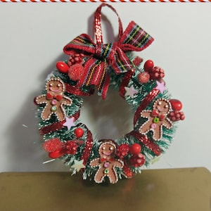 Puede incluir: Una corona de Navidad hecha de muérdago verde con cinta roja y adornos de hombre de jengibre. La corona está decorada con bayas rojas y estrellas rosas.