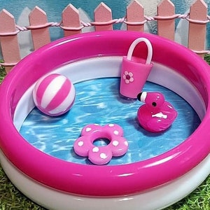 以下が含まれることがあります： 白い内側のリングが付いたピンクの空気入れ式プール。プールにはピンクのフラミンゴ、ピンクと白のビーチボール、ピンクの花、ピンクのバケツが入っています。