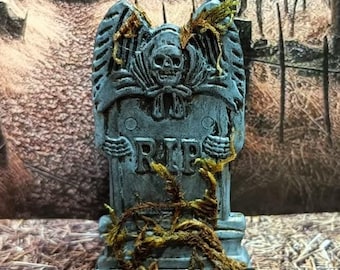 Puede incluir: Una lápida azul y gris con un diseño de calavera y huesos cruzados y la palabra "RIP" en la parte delantera. La lápida está cubierta de enredaderas y una araña negra está sentada en el suelo frente a ella.