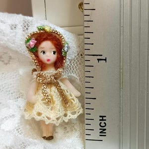 Puede incluir: Una muñeca en miniatura con cabello rojo, vestida con un vestido de ganchillo color crema y un sombrero a juego adornado con flores. La muñeca mide aproximadamente 2,5 cm de alto, con una corbata trenzada dorada alrededor del cuello. Es un objeto de colección.