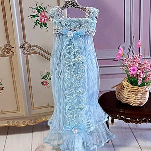 May include: A miniature light blue dress with lace detailing and a bow, displayed on a decorative hanger. The dress features a layered skirt and is designed with delicate lace trim. The overall aesthetic is delicate and feminine.