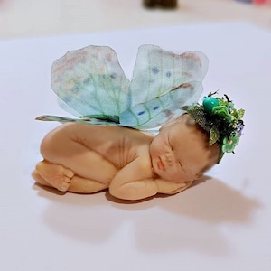 Könnte beinhalten: Eine kleine Porzellanpuppe mit Schmetterlingsflügeln und einer grünen Blumenkrone.