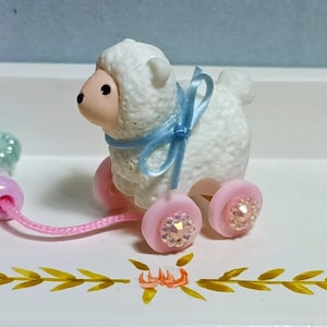 Puede incluir: Figura de cordero blanco con lazo azul claro y ruedas rosas adornadas con pedrería iridiscente. Una cuerda rosa está unida a un anillo de cuentas azul claro. Juguete infantil.