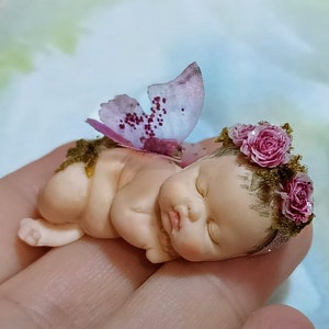 Puede incluir: Una pequeña muñeca de bebé durmiendo con una corona de flores rosas y un ala de mariposa rosa. La muñeca está hecha de porcelana y tiene un aspecto realista.