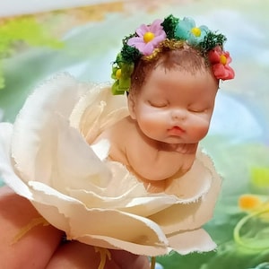 Puede incluir: Una pequeña muñeca de bebé durmiendo que lleva una corona de flores con flores rosas, azules y verdes. La muñeca está tumbada en una rosa de tela blanca.