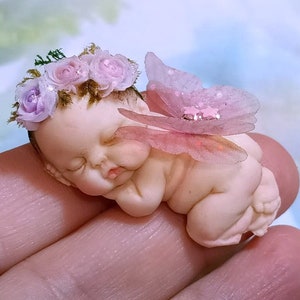 Puede incluir: Una pequeña muñeca de bebé durmiendo con alas rosas y una corona de flores. La muñeca está hecha de porcelana y tiene una apariencia delicada y realista.
