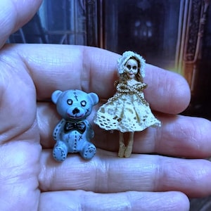 Puede incluir: Un peque&ntilde;o oso de peluche azul cosido con una pajarita negra y una mu&ntilde;eca de porcelana blanca con un vestido de encaje blanco.