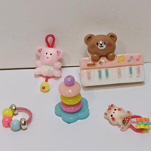 Może przedstawiać: Kolekcja miniaturowych zabawek, w tym r&oacute;żowa grzechotka w kształcie misia, brązowy miś na r&oacute;żowym pianinie, kolorowa zabawka do układania w stosy, pierścionek z koralik&oacute;w i mały biały kot z truskawką.