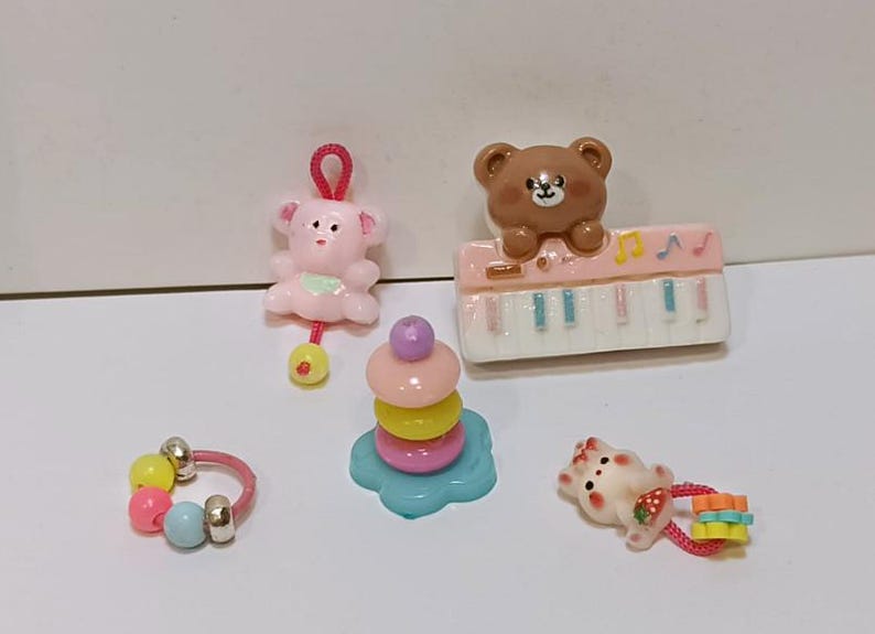 Może przedstawiać: Kolekcja miniaturowych zabawek, w tym r&oacute;żowa grzechotka w kształcie misia, brązowy miś na r&oacute;żowym pianinie, kolorowa zabawka do układania w stosy, pierścionek z koralik&oacute;w i mały biały kot z truskawką.