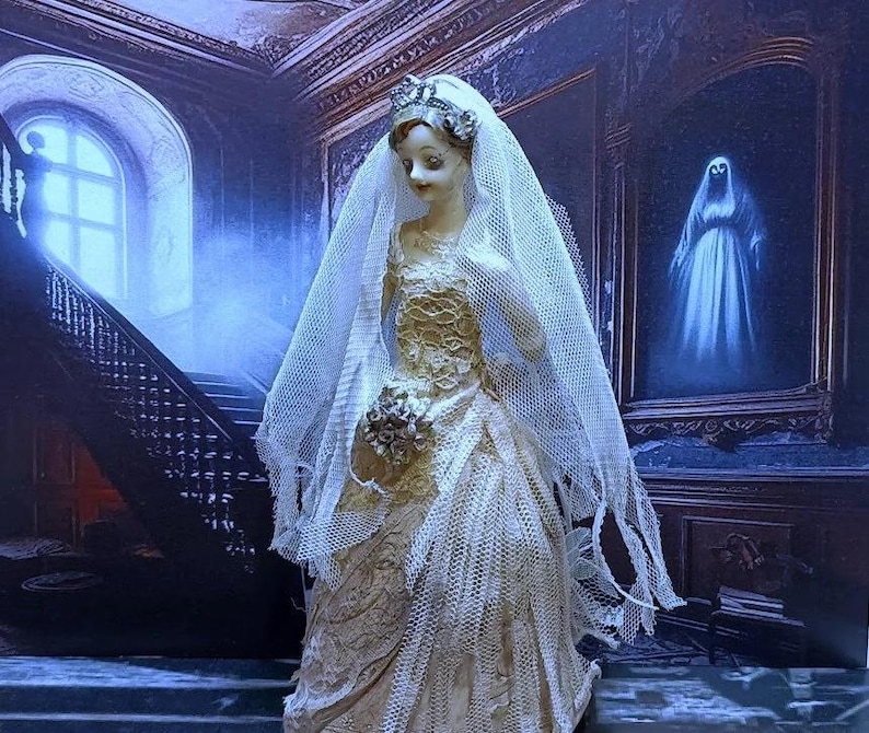 May include: A porcelain doll bride wearing a white lace wedding dress and veil. The doll is standing in a dark, gothic setting with a staircase and a painting of a ghost in the background.