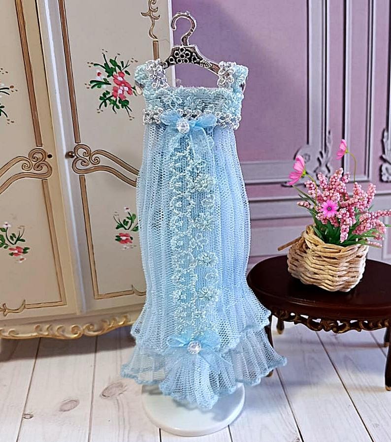 May include: A miniature light blue dress with lace detailing and a bow, displayed on a decorative hanger. The dress features a layered skirt and is designed with delicate lace trim. The overall aesthetic is delicate and feminine.