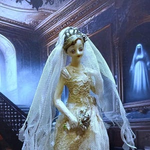 May include: A porcelain doll dressed in a white lace wedding gown and veil, holding a bouquet of flowers. The doll is standing in a dimly lit room with a staircase in the background.