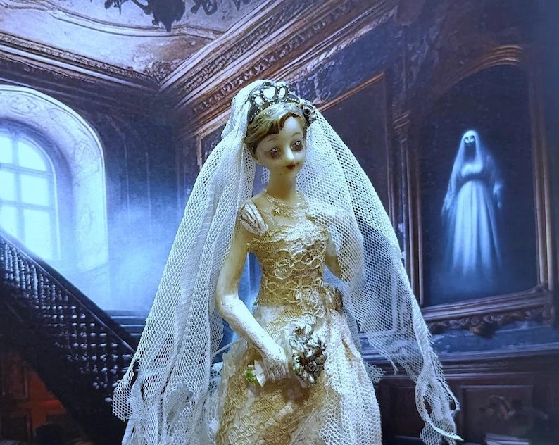 May include: A porcelain doll dressed in a white lace wedding gown and veil, holding a bouquet of flowers. The doll is standing in a dimly lit room with a staircase in the background.