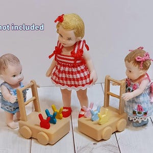 May include: Two miniature wooden pull-along carts, each featuring four wheels and a handle, with three small colorful rabbit-shaped figures attached to the top of each cart. The text "Dolls not included" is visible in the upper left corner.