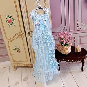 May include: A light blue doll dress with lace detailing, hanging on a decorative hanger. The dress has a ruffled hem and is displayed in front of a vintage-style cabinet. A small table with a flower arrangement is in the background.