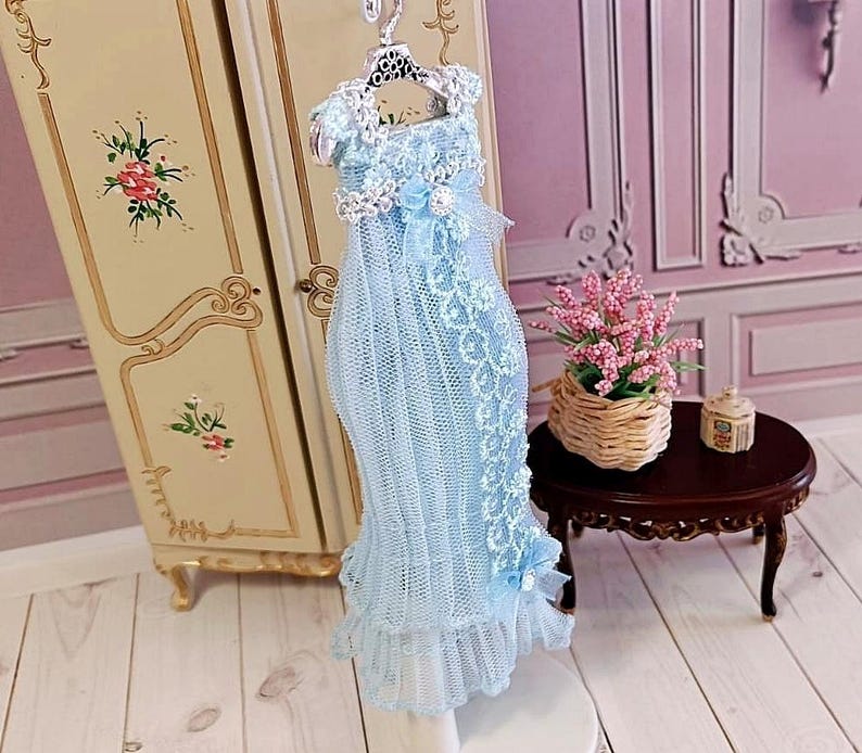 May include: A light blue doll dress with lace detailing, hanging on a decorative hanger. The dress has a ruffled hem and is displayed in front of a vintage-style cabinet. A small table with a flower arrangement is in the background.