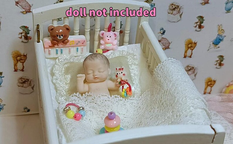 Może przedstawiać: Miniaturowe białe ł&oacute;żeczko ze śpiącą lalką-niemowlęciem, misiem i innymi zabawkami. Ł&oacute;żeczko wypełnione jest białą koronkową kołdrą i poduszką. Tło przedstawia tapetę z postaciami z kresk&oacute;wek. U g&oacute;ry widnieje napis "lalka nie jest dołączona."