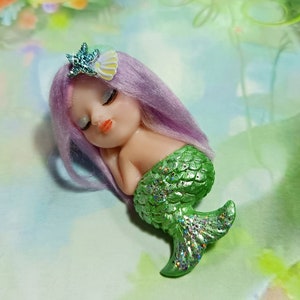 May include: A small, sleeping mermaid figurine with purple hair and a green, glittery tail. She is wearing a small, blue starfish hair clip.