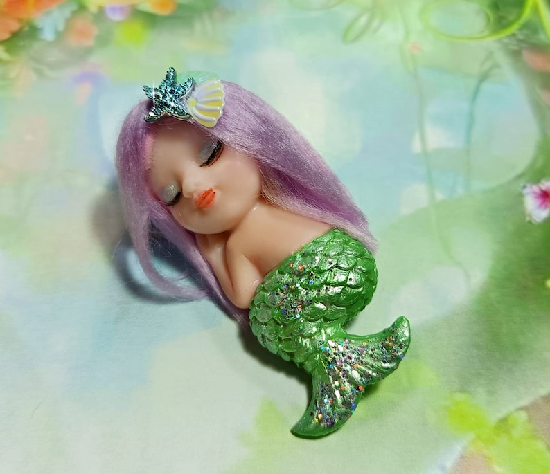 May include: A small, sleeping mermaid figurine with purple hair and a green, glittery tail. She is wearing a small, blue starfish hair clip.