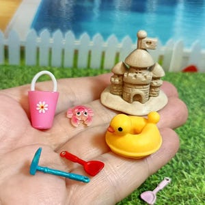 May include: Miniature beach-themed toys including a pink bucket with a daisy, a sandcastle, a yellow duck float, a crab, and blue and red shovels. These small toys are perfect for dollhouses or craft projects.