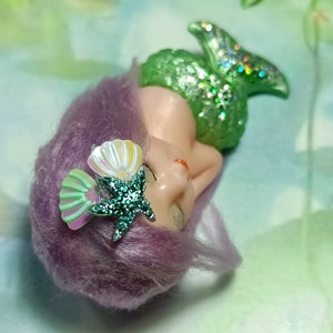 May include: A small mermaid figurine with a green glittery tail, purple hair, and a seashell and starfish hair accessory.