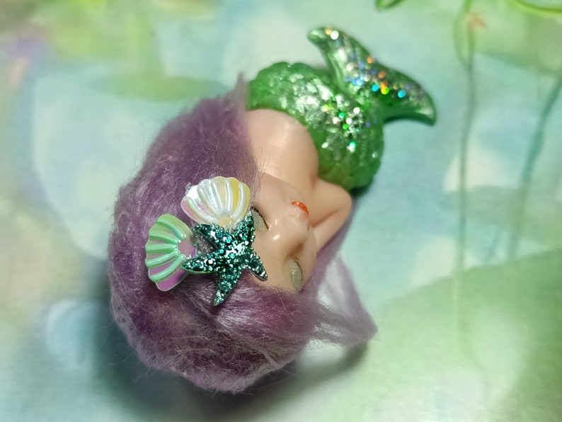 May include: A small mermaid figurine with a green glittery tail, purple hair, and a seashell and starfish hair accessory.