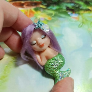 May include: A small, handmade mermaid figurine with purple hair, closed eyes, and a green glittery tail. The mermaid is wearing a starfish and seashell hair accessory.
