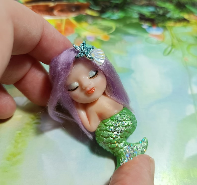 May include: A small, handmade mermaid figurine with purple hair, closed eyes, and a green glittery tail. The mermaid is wearing a starfish and seashell hair accessory.