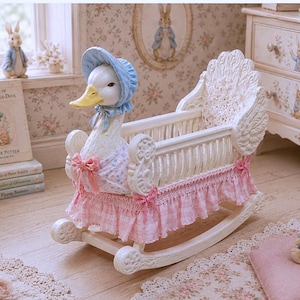 Puede incluir: Una cuna mecedora decorativa blanca con una cabeza de pato esculpida en la parte delantera que lleva un gorro azul. La cuna tiene una falda de tela a cuadros rosas y blancos con lazos rosas, un marco de listones blancos y un cabecero alado.