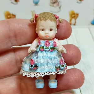 Puede incluir: Una muñeca en miniatura con cabello castaño claro peinado en dos coletas con lazos rosas. La muñeca viste un vestido de cuadros azul claro y blanco con bordados florales rosas en el cuello y el dobladillo, rematado con encaje blanco y zapatos azules.