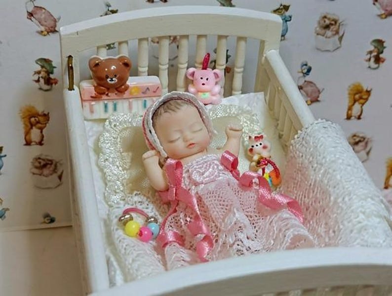 Może przedstawiać: Miniaturowa lalka w białym ł&oacute;żeczku, ubrana w r&oacute;żową sukienkę i czepek. Ł&oacute;żeczko ma białą ramę i jest ozdobione koronkowym kocykiem i małymi zabawkami, w tym misiem i r&oacute;żowym misiem. Tło przedstawia tapetę z ilustracjami zwierząt.