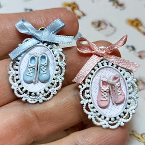 Puede incluir: Dos colgantes ovalados en miniatura con diseños de zapatos de bebé. Uno presenta zapatos azul claro y un lazo a juego, el otro tiene zapatos rosas y un lazo rosa. Ambos están en marcos blancos ornamentados.