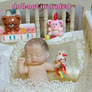 Może przedstawiać: Miniaturowa lalka w białym ł&oacute;żeczku z koronkową lam&oacute;wką. Ł&oacute;żeczko ma białe wezgłowie i podn&oacute;żek. Lalka trzyma małą zabawkę. Ł&oacute;żeczko jest ozdobione miniaturowymi zabawkami, w tym misiem i r&oacute;żowym misiem.