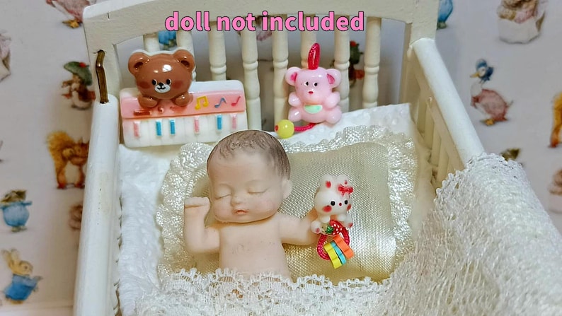 Może przedstawiać: Miniaturowa lalka w białym ł&oacute;żeczku z koronkową lam&oacute;wką. Ł&oacute;żeczko ma białe wezgłowie i podn&oacute;żek. Lalka trzyma małą zabawkę. Ł&oacute;żeczko jest ozdobione miniaturowymi zabawkami, w tym misiem i r&oacute;żowym misiem.