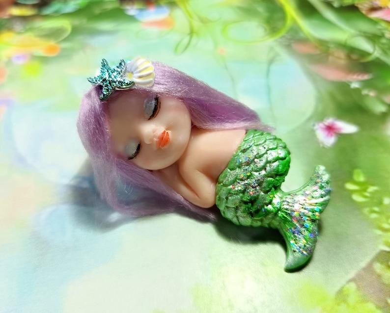May include: A small, sleeping mermaid doll with purple hair and a green, glittery tail. The doll is wearing a starfish and seashell hair accessory.