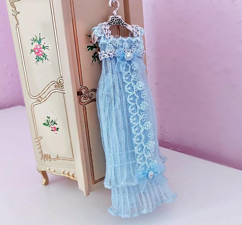 May include: A miniature light blue dress with lace detailing, hanging on a decorative hanger. The dress features a layered skirt and a bow at the waist. The dress is displayed next to a small, ornate cabinet.
