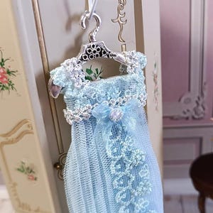 May include: A miniature light blue dress with intricate lace detailing, hanging from a silver-colored hanger. The dress features a delicate bow and a pearl-like embellishment. The background includes a glimpse of a decorative cabinet and a small table, suggesting a dollhouse or miniature setting.