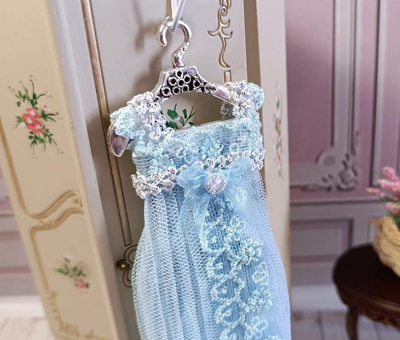 May include: A miniature light blue dress with intricate lace detailing, hanging from a silver-colored hanger. The dress features a delicate bow and a pearl-like embellishment. The background includes a glimpse of a decorative cabinet and a small table, suggesting a dollhouse or miniature setting.