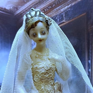 May include: A porcelain doll bride wearing a white veil and a gold and white dress. The doll has a crown on her head and is standing in front of a window with a staircase leading up to it.