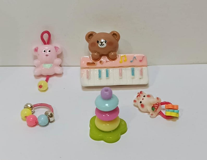 Może przedstawiać: Kolekcja miniaturowych zabawek, w tym r&oacute;żowa zabawka do ciągnięcia w kształcie misia, brązowy miś na r&oacute;żowym pianinie, kolorowa zabawka do układania w stos, grzechotka i mały miś z truskawką. Zabawki są w r&oacute;żnych kolorach.