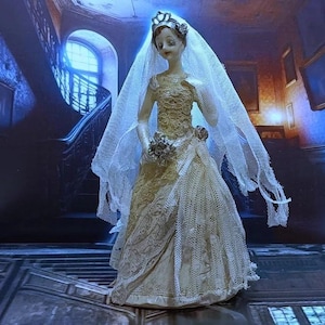 May include: A porcelain doll bride figurine wearing a white lace wedding dress and veil. The doll is standing on a wooden floor with a staircase in the background.